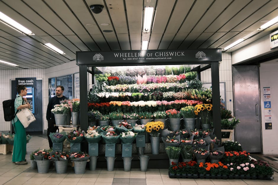 A vibrant flower display at Wheeler of Chiswick within a busy underground station. The arrangement features a variety of fresh flowers including yellow sunflowers, pink roses, red carnations, white li