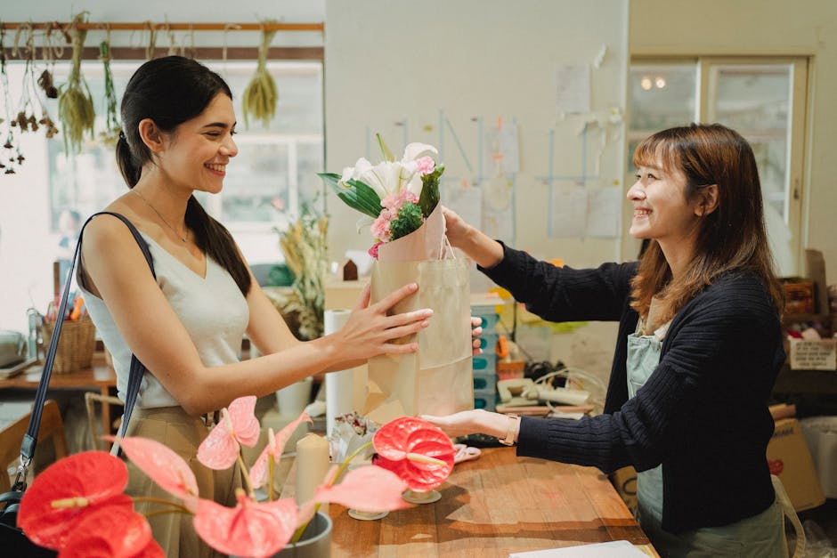 A floral arrangement in a light pink paper wrapping featuring white and soft pink roses, green foliage, and other pastel-colored blooms, being handed over at a florist shop counter. A smiling female s