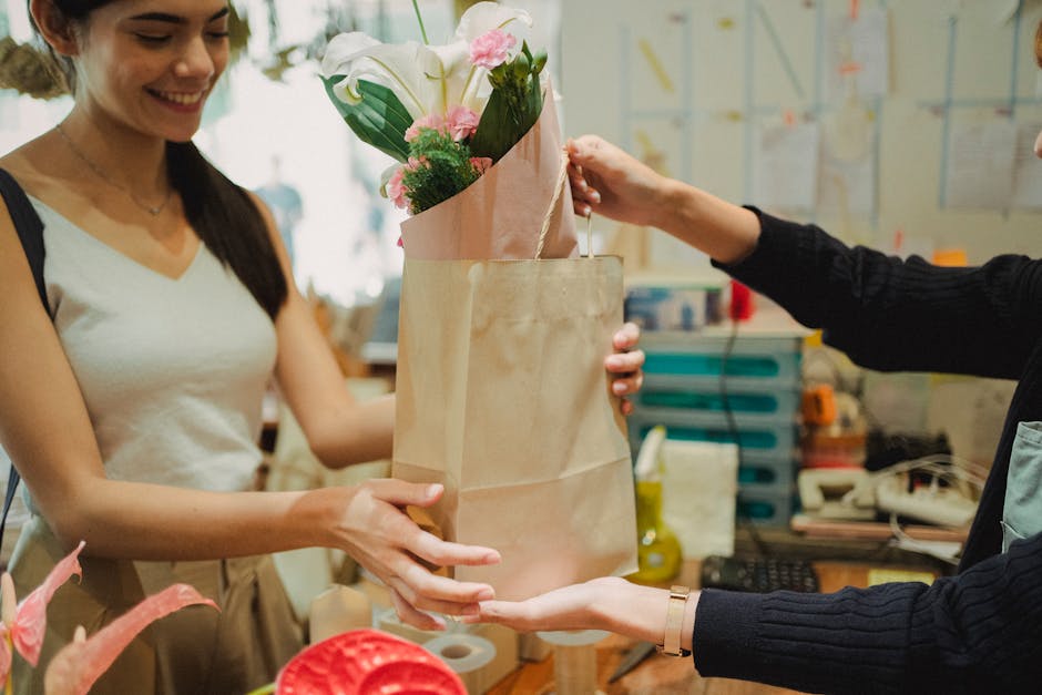 A woman with long dark hair, wearing a white sleeveless top, is smiling while receiving a bouquet of fresh flowers from a florist at a floral shop. The bouquet, wrapped in pink paper, includes elegant